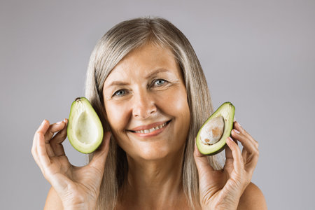 Portrait of pleasant mature woman posing in studio with halves of green ripe avocado in hands....