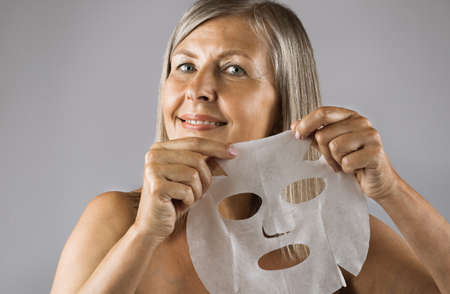 Portrait of beautiful senior woman smiling on camera with cotton mask sheet in hands. Isolated over grey studio background. Concept of face care and beauty treatment.の写真素材