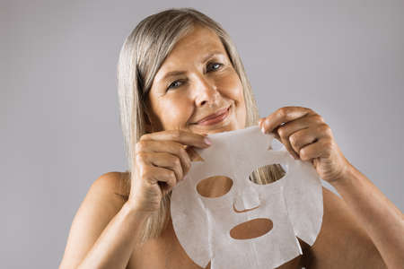 Portrait of beautiful senior woman smiling on camera with cotton mask sheet in hands. Isolated over grey studio background. Concept of face care and beauty treatment.の写真素材
