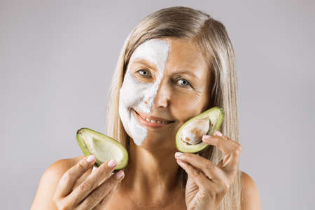Smiling senior woman holding avocado near her face with white clay mask on half face against grey background. Concept of people, skin care and organic cosmetic.の写真素材