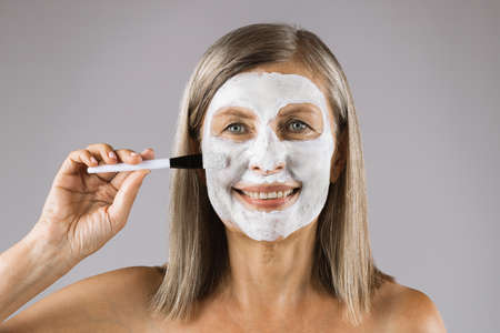 Attractive mature lady smiling and looking at camera with white clay mask on face. Caucasian woman using brush for applying cosmetic against grey background.の写真素材