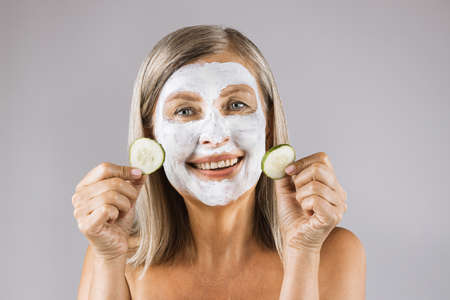 Portrait of positive caucasian woman holding slices of cucumber in hands while wearing white cosmetic mask on face. Isolated over grey studio background.の写真素材