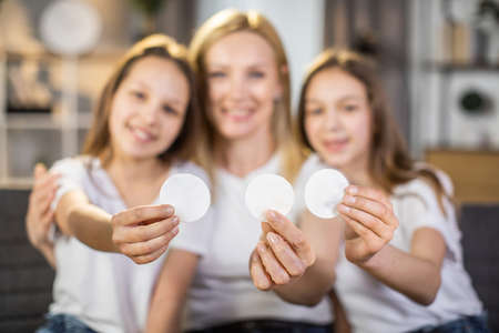 Blur background of two caucasian sisters sitting in embrace with charming mother hand holding cotton pads in hands. Healthy beauty routine at home.の写真素材