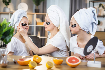 Two sisters and caucasian woman with bath towel on head sitting at desk and using black hydrogel eye patches. Caring mother teaching her teen daughters beauty routine for healthy and fresh skin.の写真素材