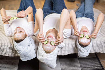 Attractive caucasian woman lying on couch with her pretty daughters, having mask on face and slices of cucumber on eyes. Domestic beauty procedures and skin care concept.の写真素材