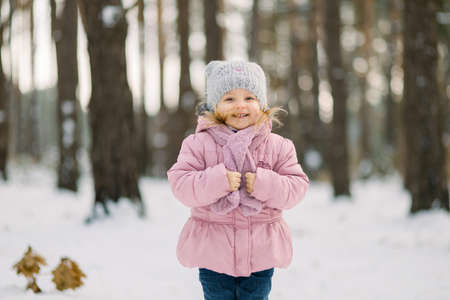 Winter in the forest. Cute dreamy little girl in a pink coat standing in snowy forest with trees background and looking on camera. Smiling kid walking in frosty winter day in forest.の写真素材