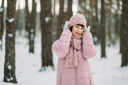Outdoor winter portrait of pretty young woman wearing pink coat and stylish knitted hat and scarf, standing in beautiful snowy forest in front of pine trees and enjoying walk on frosty dayの写真素材