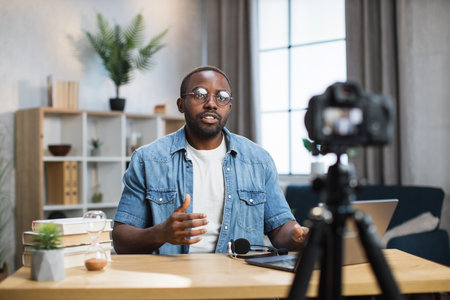 African american man in denim shirt and eyeglasses sitting at table, gesturing and talking while recording video on camera. Male blogger doing live stream in his social network.の写真素材