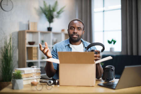 African american man in casual wear taking modern headphones for listening to music from paper box while sitting at the table at home. Concept of technology and blogging.の写真素材