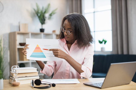 Smart female teacher in eyeglasses pointing on diagram during video lesson from home. African...