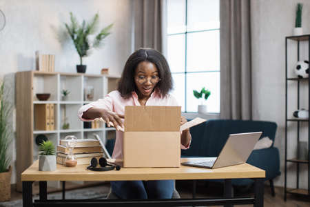 Excited african american woman sitting at desk and opening parcel box. Female buyer checking ordered goods from delivery service. Concept of people and purchases.の写真素材
