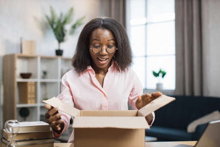 Attractive african woman with surprised facial expression looking inside paper box. Happy young lady in eyeglasses and pink shirt unpacking parcel.の写真素材