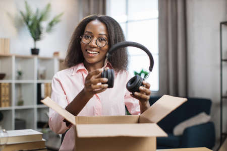 African american woman smiling and looking at camera while unpacking parcel box with new headphones. Concept of shopping online and good delivery.の写真素材