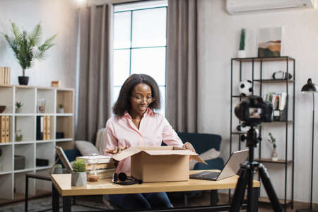 Happy african woman doing live stream while unpacking parcel box at home. Popular female blogger using modern video camera for work.の写真素材