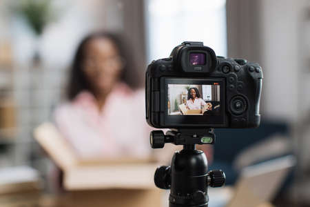 African american woman unboxing parcel with ordered goods while sitting at home and recording video blog. Female influencer using modern digital camera for creating new content.の写真素材
