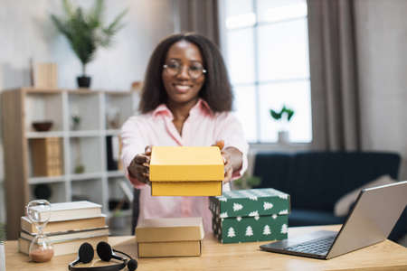 Smiling african woman in casual wear sitting at desk and holding colorful gift boxes in hands. Famous female influencer opening festive presents made by worldwide companies.の写真素材
