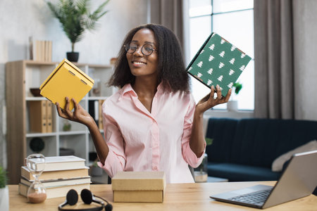 Beautiful african woman in eyeglasses holding colorful gift box in hands while sitting at desk and...
