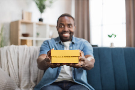 African american man in denim shirt holding yellow gift box while sitting on cozy sofa. Positive...