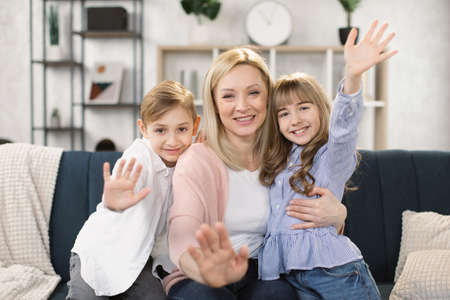 Happy mom and two little sibling kids enjoying leisure at home together, relaxing on couch, hugging, waving their hands. Loving mother embracing sweet children with care, affection. Family conceptの写真素材