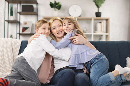 Happy little kids sit on couch hug cuddle excited young mom or nanny show love and care, overjoyed small children relax with mother embrace share sweet tender family moment in living room at homeの写真素材