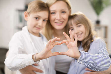Cute little daughter with son and mother join hands in shape of heart as concept of mom and kids love care support, smiling mum and her kids looking at camera posing together for headshot portraitの写真素材