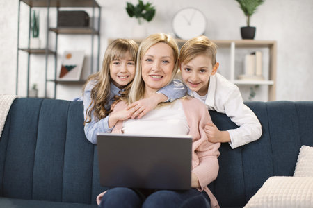 Happy young caucasian mother relax on comfortable couch with little kids have fun watch video on...