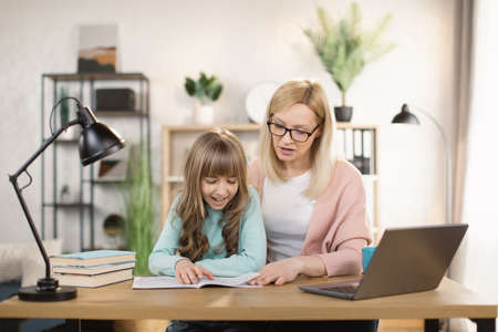 Young caucasian mother helping daughter remote studying at home explaining doing homework together. Parent mom teaching teenage school child distance learning online virtual class on computer.の写真素材
