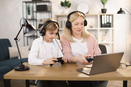 Happy woman and son play video games sitting at a table in the living room at home. A gambling boy and his mother are playing with the game console joysticks in their hands. A happy family.の写真素材