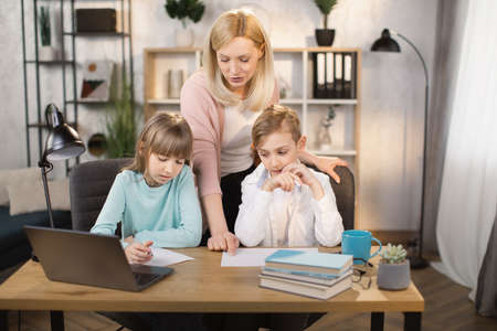 Dughter and son doing school lessons together looking at a laptop monitor. Mother stands behind and watches and shows the task on paper.の写真素材
