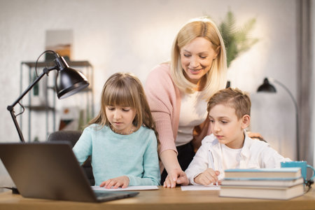 Dughter and son doing school lessons together looking at a laptop monitor. Mother stands behind and...