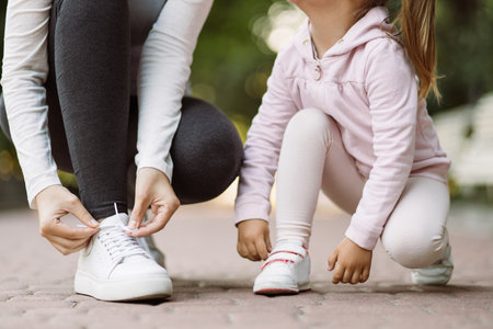 Family walk and fit sport training. Cropped close up shot of legs of sporty mother and little daughter adjusting sneakers, sitting on the jogging track in the parkの写真素材