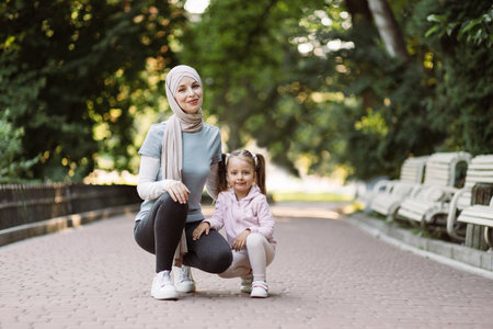 Positive good-looking young mother in hijab, posing together with her little daughter, ready to do physical activity outdoors in the green park. Family, sport and recreation conceptの写真素材