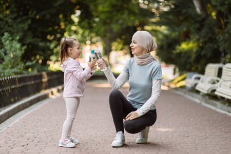 Likable pretty young Muslim lady in hijab and sportswear posing together with her cute little daughter and clinking water bottles on camera. Family sport workout in parkの写真素材