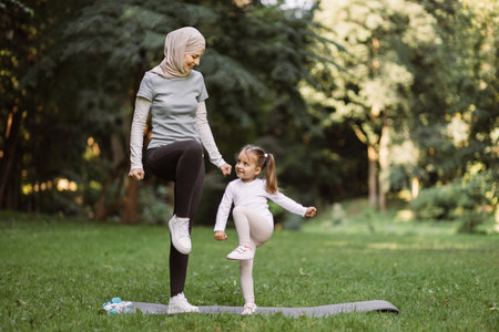 Yoga and stretching exercises of mom with child outdoors. Likable young Muslim mother and her cute daughter, having outdoor workout in the park, standing on yoga mat and doing high stepping exerciseの写真素材