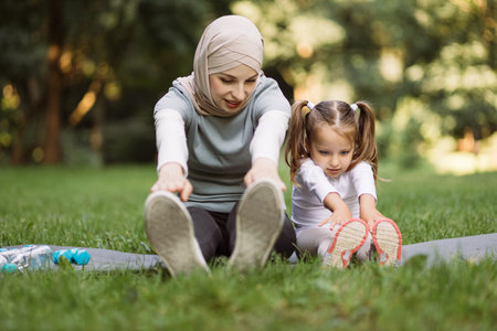 Vitality and healthy lifestyle concept. Cute active preschool girl and her slim sportive Muslim mom are doing stretching exercises in green summer park sitting on gray matの写真素材