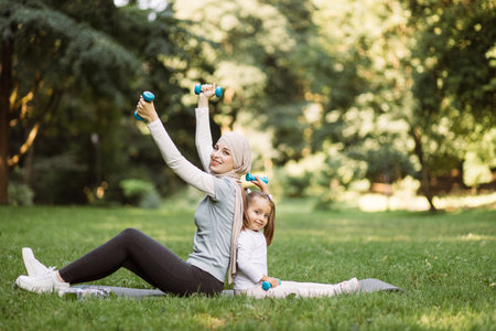 Family sport activity outdoors. Pretty sporty fit Muslim Arabian woman working out with dumbbells together with her cute 4 years old daughter, sitting on yoga mat in the summer parkの写真素材