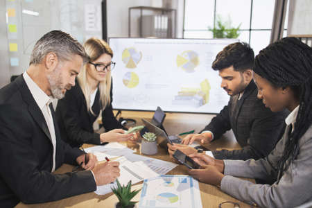 Diverse group of multiracial business people in the conference room with big TV screen, cooperating at the table, looking at papers with financial charts and statistics. Teamwork concept.の写真素材