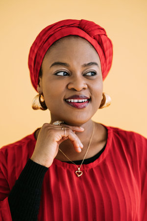 People, races and ethnic beauty concept. Close up portrait of charming African woman with red traditional shawl on her head, jewerly and bright make up, posing on yellow background looking awayの写真素材