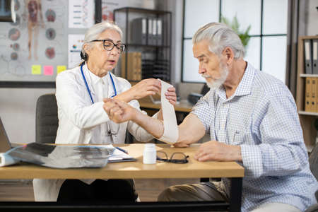 Mature female therapist in eyeglasses and white lab coat wrapping bandage on arm of senior man. Qualified doctor taking care of injury of male patient at medical cabinet.の写真素材