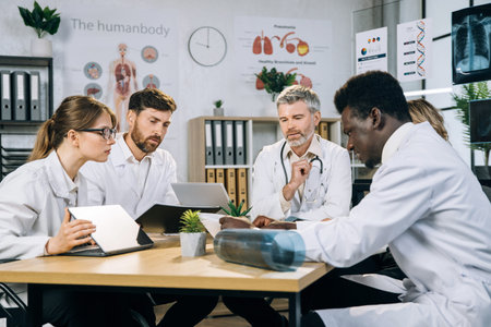 International group of five qualified doctors sitting together at table and using modern gadgets...