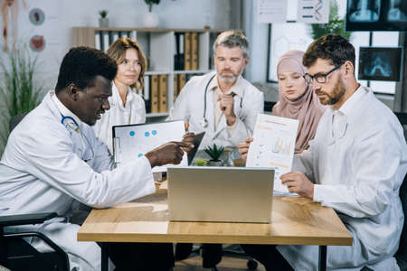 Five multi ethnic medical specialists showing various charts and graphs during video call on modern laptop. Group of competent doctors having online conference at office.の写真素材