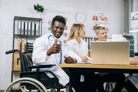 African wheelchair user in white lab coat smiling and looking at camera with thumb up while his...