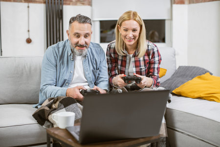 Joyful caucasian couple using wireless joysticks and laptop for playing video games at home. Mature...