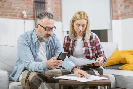 Mature caucasian couple in casual wear checking their bills at home while sitting together on couch with documents and smartphone. Domestic finances concept.の写真素材