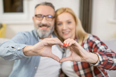 Caucasian married couple in casual attire forming heart with their fingers while sitting on comfy couch. Focus on peoples hands making love symbol.の写真素材