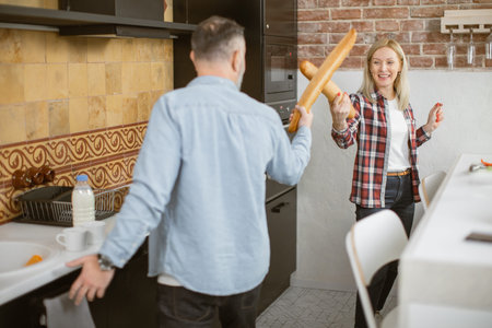 Smiling mature family in casual clothes having playful fight with fresh long baguettes on modern domestic kitchen. Happy married couple in casual wear having fun together at home.の写真素材