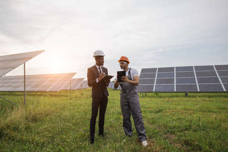 African american man in suit and indian man in uniform standing among solar plant and checking working process. People using digital tablet and clipboard during examination of panels.の写真素材