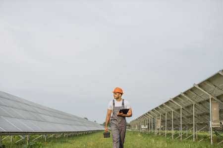 Indian man in uniform and helmet examining work of solar panels while walking on station. Competent...