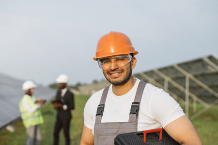 Smiling indian foreman in orange helmet and safety glasses looking at camera with two colleagues...