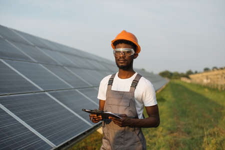 Portrait of african american man in safety glasses and orange helmet holding clipboard while standing on field with solar panels. Concept service work and alternative energy.の写真素材